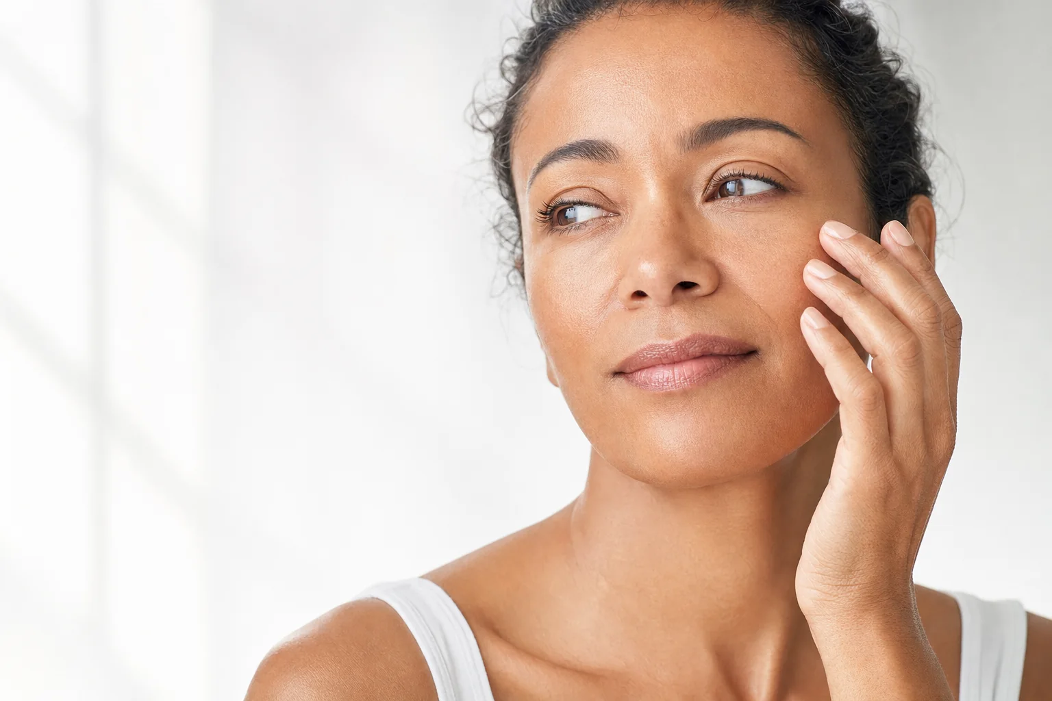A woman with warm brown skin touching her cheekbone in soft window light