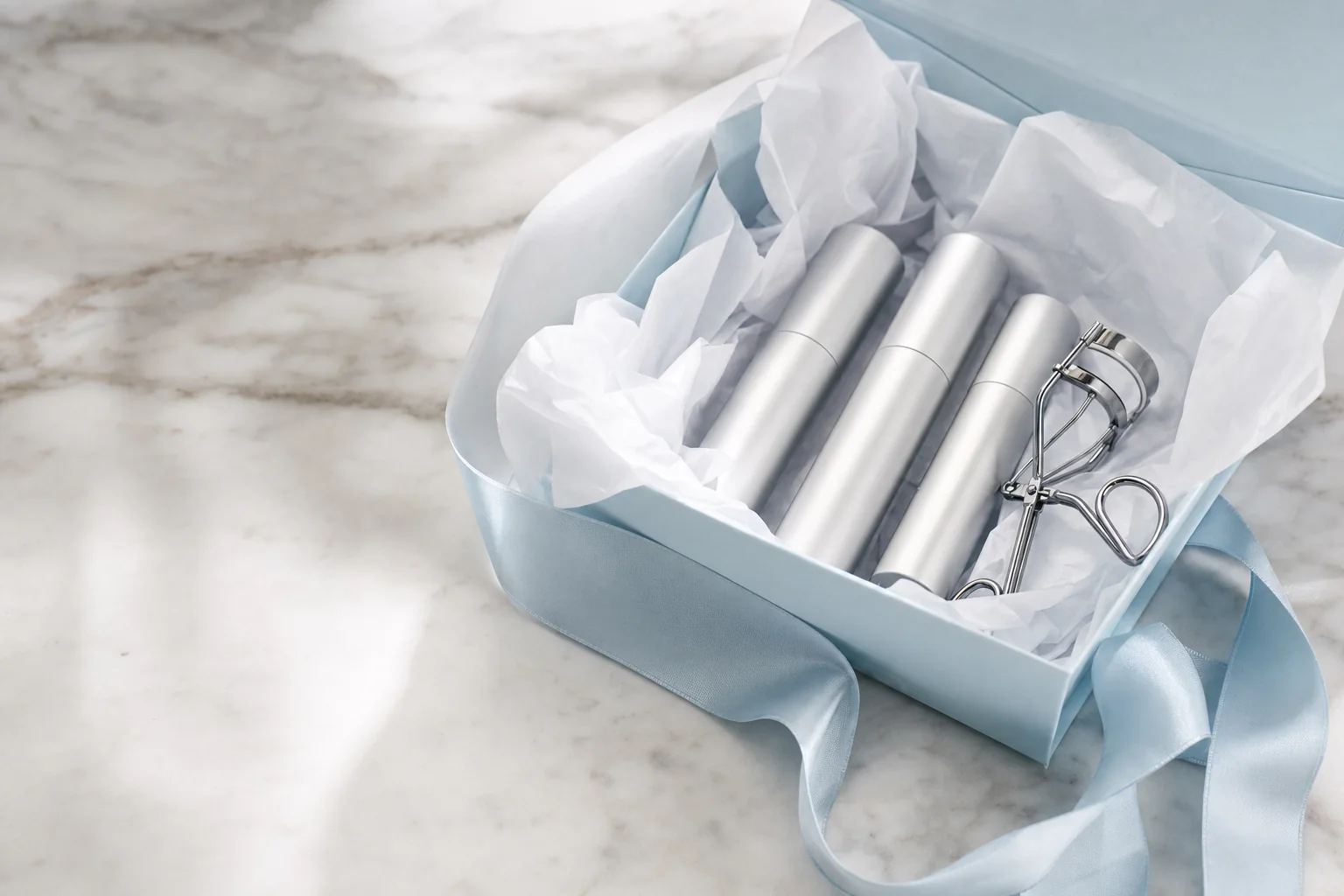 An open pale blue gift box of slim silver tubes and an eyelash curler