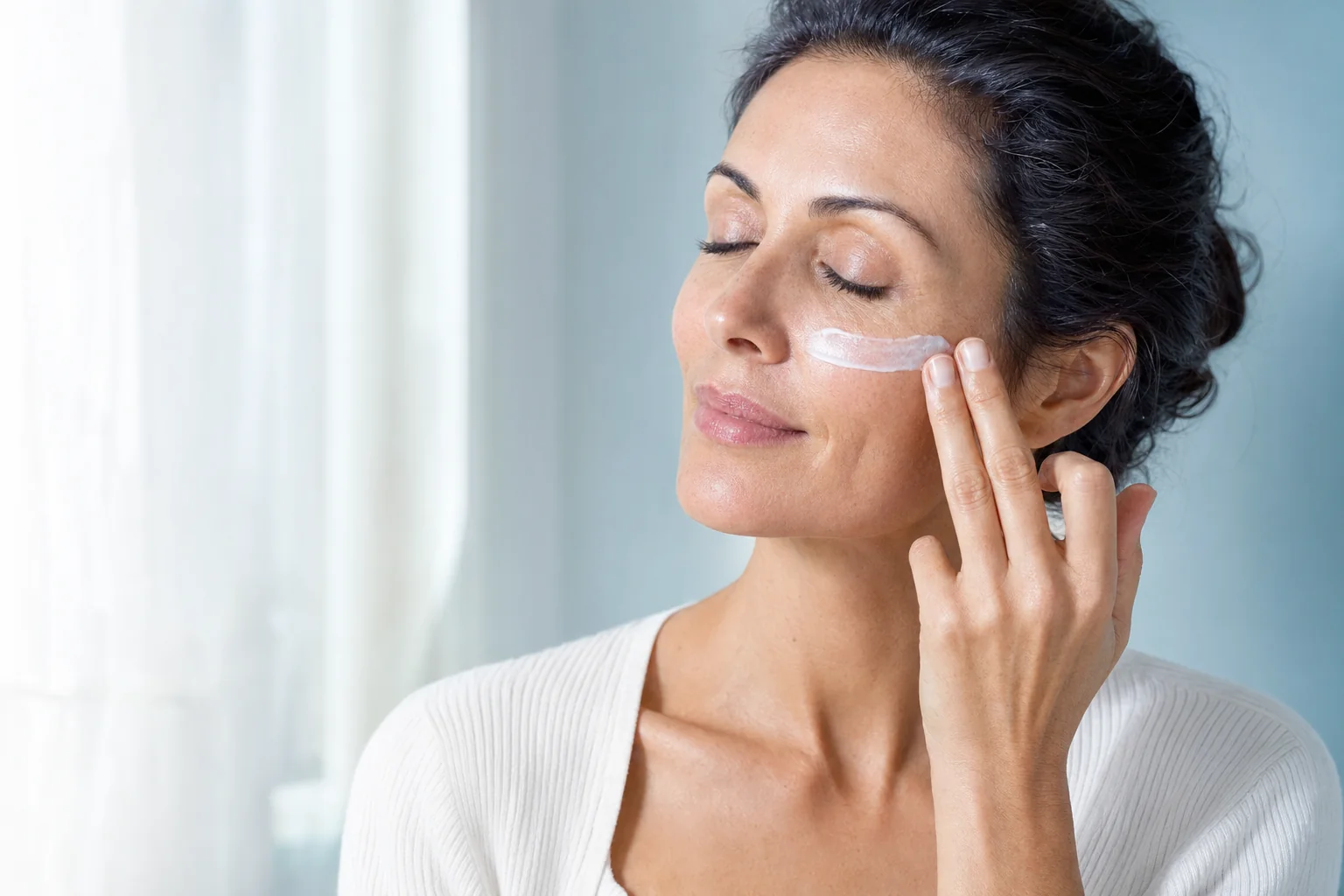 A woman smoothing a streak of white sunscreen across her cheekbone beside a bright, curtained window